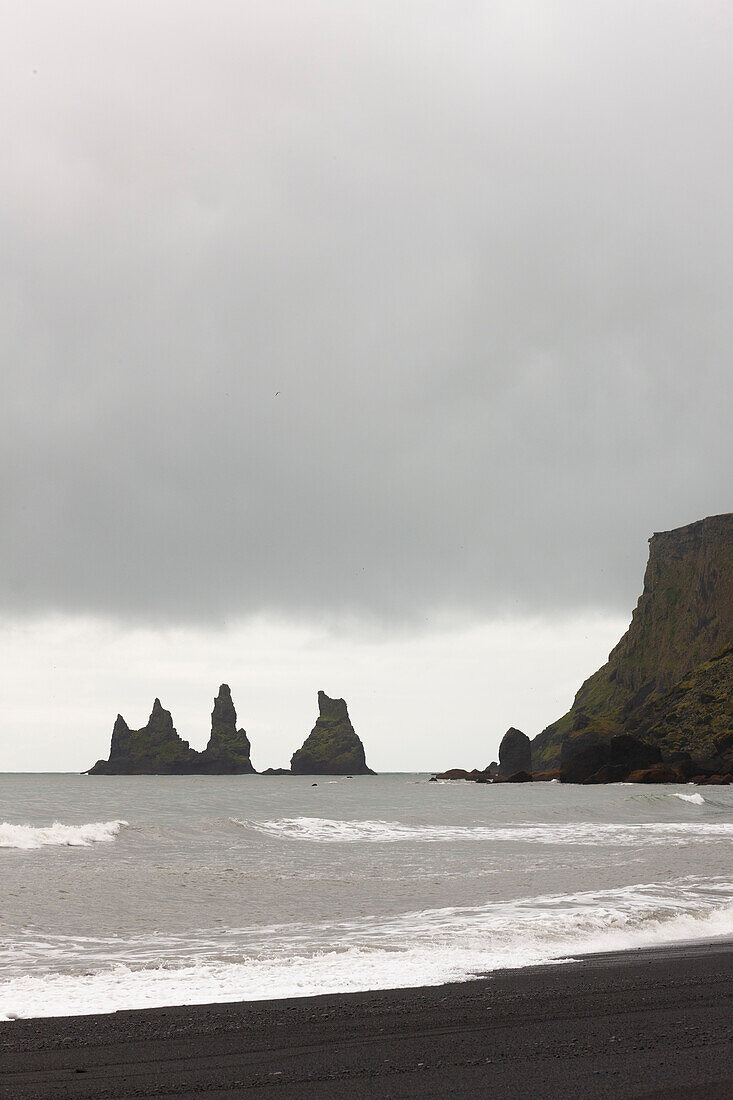 Schwarzer Sandstrand mit Reynisdrangar-Basaltfelsen und Klippen im Hintergrund an einem bewölkten Tag, in der Nähe von Vik i Myrdal, Island