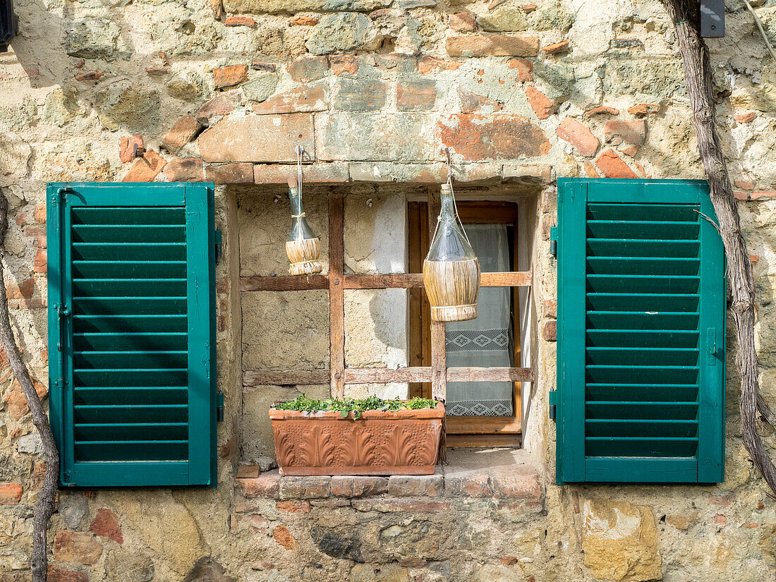 Italien, Chianti, Monteriggioni. Vergittertes Fenster mit Fensterläden, Weinflaschen und Pflanzkübel in der Hügelstadt.