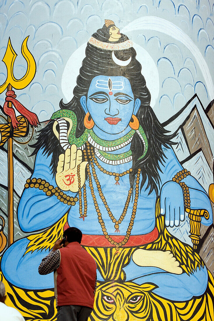 Guy using mobile phone passing mural of Shiva, one of the most famous Hindu Gods/deities painted on a tower at Dashashwamedh Ghat the most famous and central bathing ghat. The culture of Varanasi is closely associated with the River Ganges and the river's