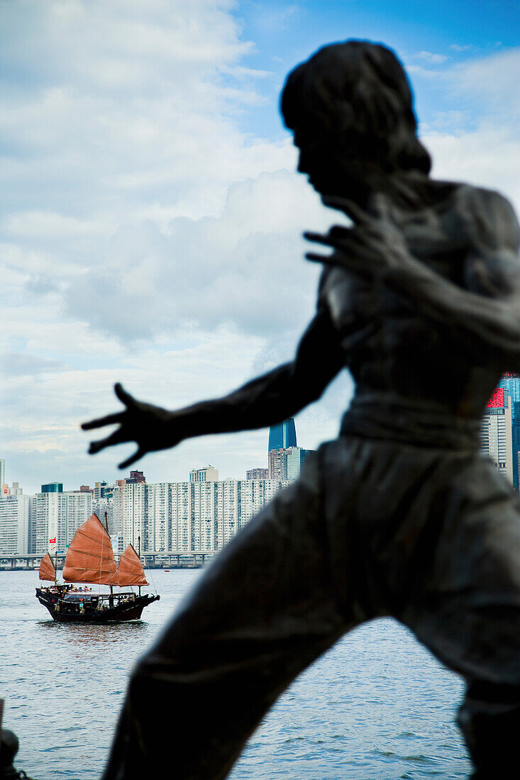 Traditionelles chinesisches Dschunkenboot im Hintergrund. Bruce Lee Memorial Statue, Blick auf Victoria Harbour, Hong Kong Island Skyline, von Tsim Sha Tsui, Kowloon; Tsim Sha Tsui, Kowloon, Hong Kong, China