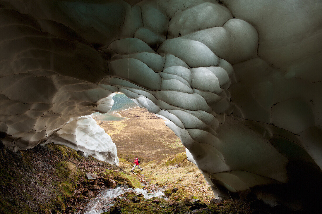 Wanderer, der über einen kleinen Bach springt, der aus einer Eishöhle kommt, Gleann Einich bei Aviemore, Highlands, Schottland, Großbritannien