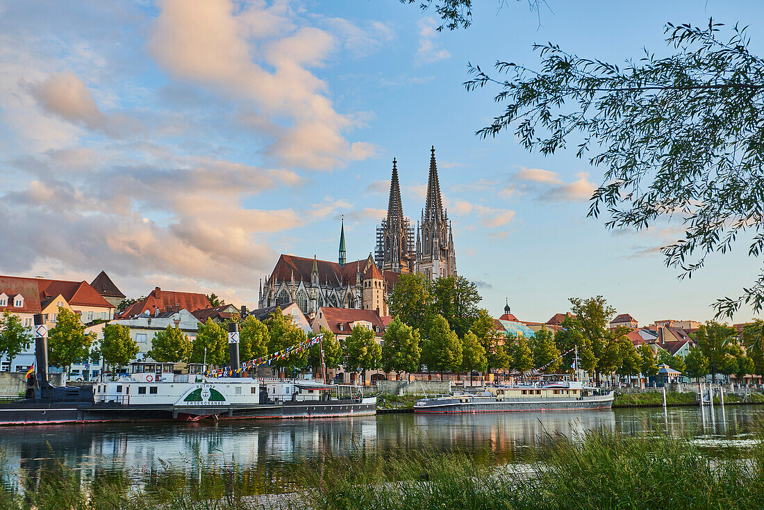 Outlook over the Danube River with the Gothic St Peter's Cathedral from the Marc?-Aurel-shore in the Old Town of Regensburg with historical steamboats moored along the riverfront at sunset; Regensburg, Bavaria, Germany