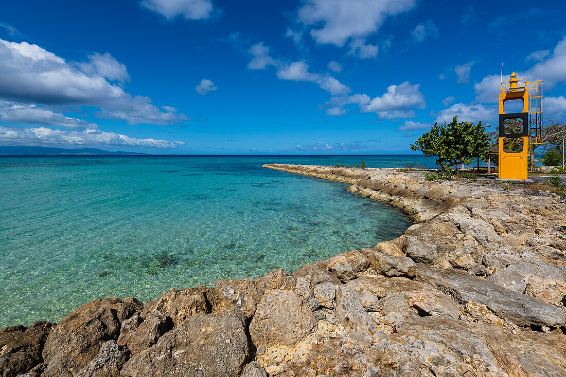 Blue sky and turquoise water of the Caribbean Sea and a small, yellow lighthouse with a black horizontal band, at the end of the rocky waterfront in Port-Louis on Grande-Terre; Guadeloupe, French West Indies