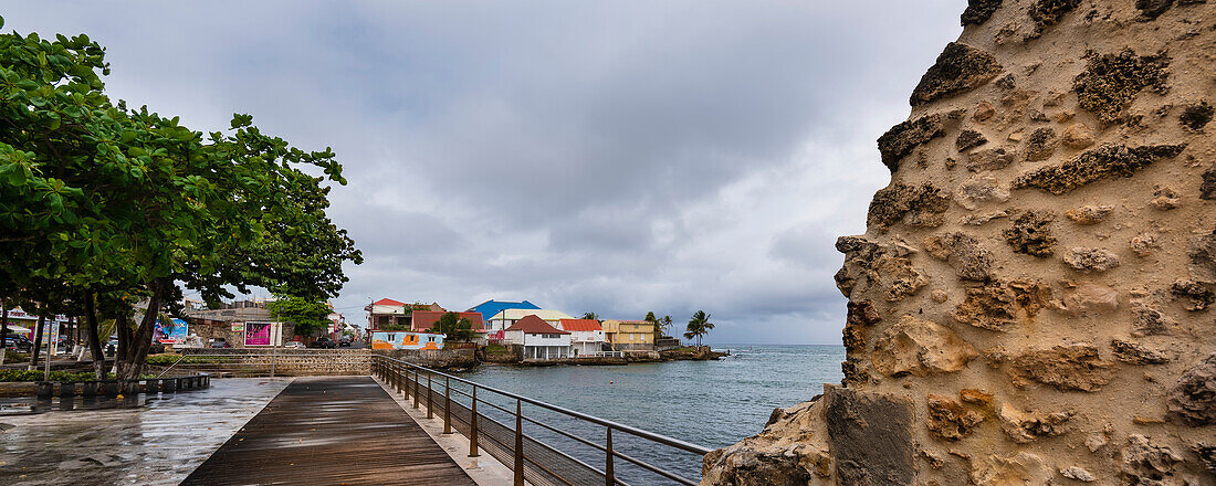 Boardwalk along the waterfront of the town of Le Moule on a cloudy day with the Wizosky ruins of the old lemonade factory on the shore of the Caribbean Sea, Grande-Terre; Guadeloupe, French West Indies