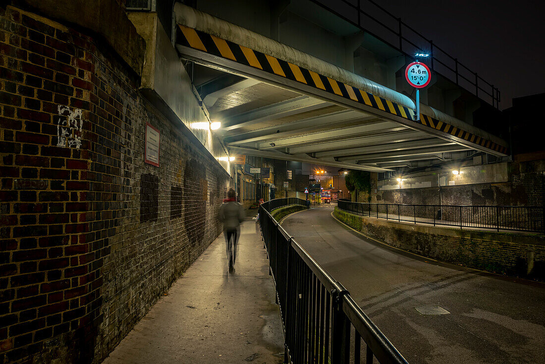 Pedestrian walking along sidewalk under a street bridge at night around Haggerston; London, England