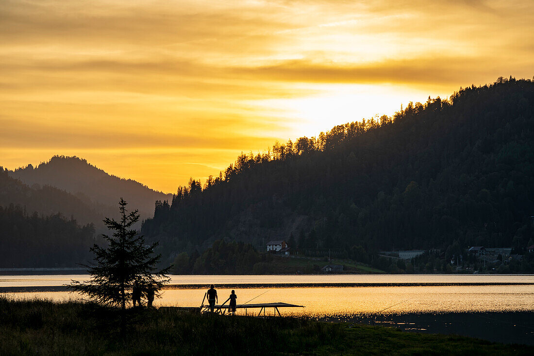 Morgendämmerung über den Karpaten in der Nähe von Tasuleasa Sozial-NGO für den Wanderweg Via Transilvanica durch Siebenbürgen mit Silhouette von Menschen beim Angeln am sonnenbeschienenen See; Siebenbürgen, Rumänien