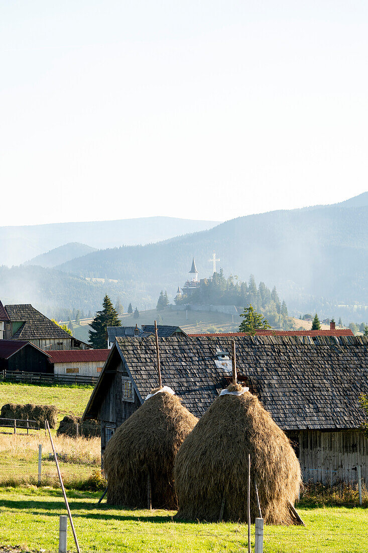 Bauernhäuser und traditionelle rumänische Heuschober mit einem Kirchturm und einem Kreuz im Hintergrund in der ländlichen Umgebung von Siebenbürgen; Siebenbürgen, Rumänien