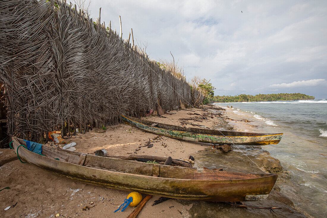 Zwei gestrandete Kanus und eine Mauer aus geflochtenen Sagopalmen (Cycas revoluta), die als Windschutz für ein Dorf an der Luvküste von Tuam Island, einer der Siassi-Inseln in der Bismarcksee vor der Nordküste Papua-Neuguineas, errichtet wurde; Siassi, Vitiaz Strait, Papua-Neuguinea