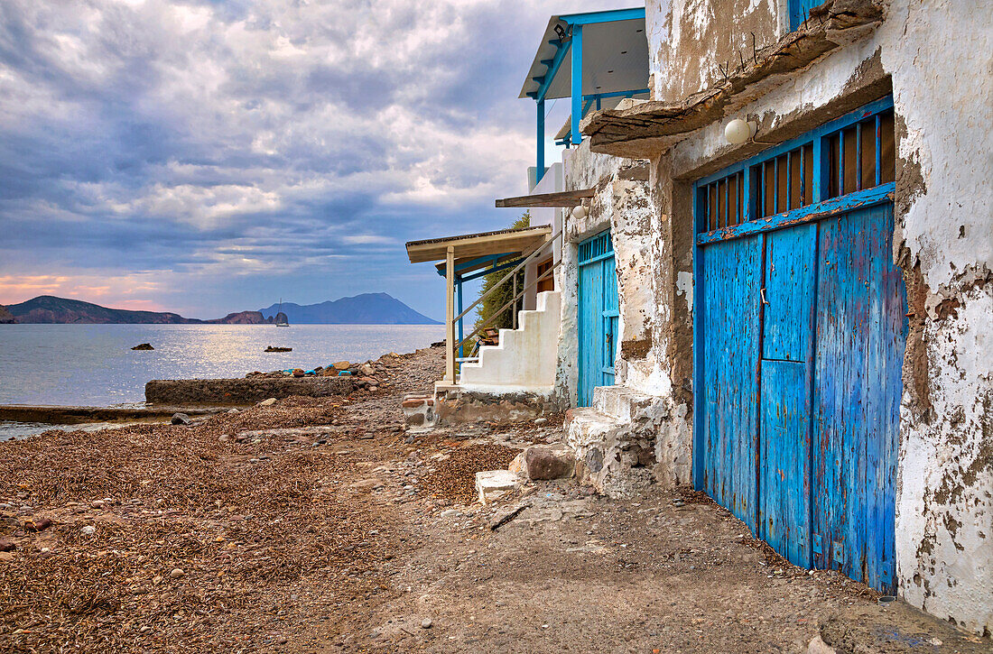 Dilapidated stone buildings with painted, blue wooden doors next to the shore with mountains on the horizon at twilight