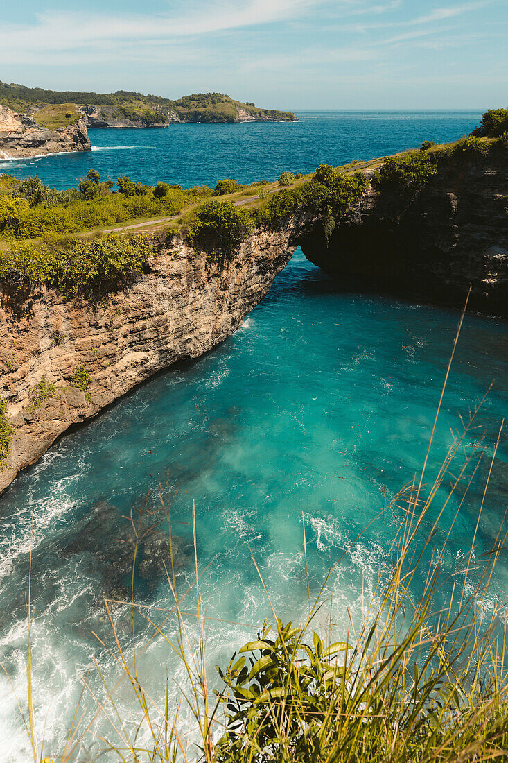Grün bewachsener Meeresbogen, Felsformation mit türkisfarbenem Wasser und Meeresbrandung entlang der felsigen Küstenlinie der Nusa Inseln; Nusa Penida, Provinz Bali, Indonesien