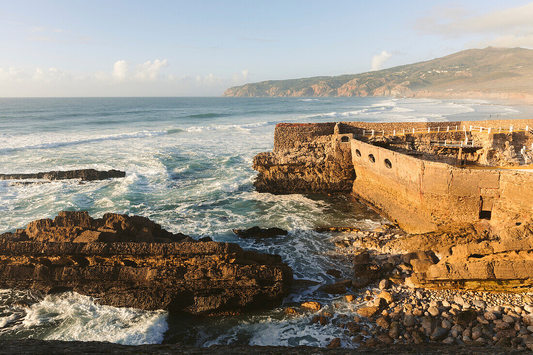Wellen des Atlantischen Ozeans am Ufer des Praia do Guincho bei Cascais mit dem Außenpool des Hotels Estalagem Muchaox, der in die Steinmauer-Ruinen einer alten Festung gebaut wurde; Praia do Guincho, Cascais, Lison, Portugal