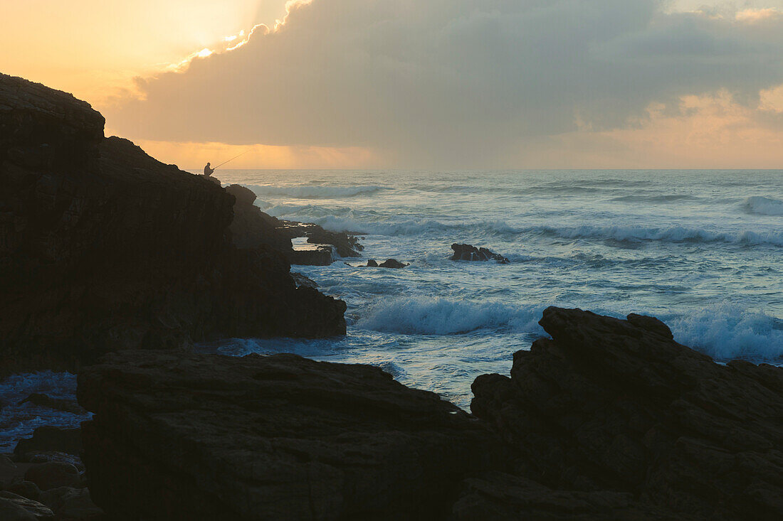 Silhouette of man surf fishing along a rugged coastline at sunset; Praia do Guincho, Cascais, Portugal