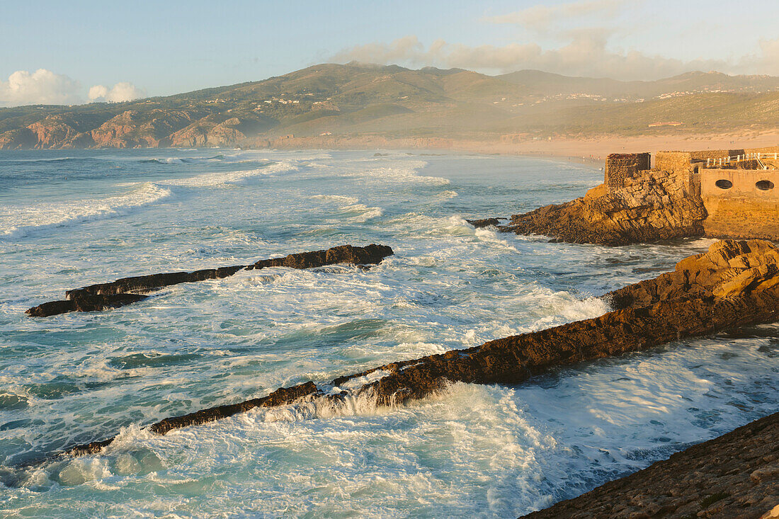 Wellen des Atlantischen Ozeans entlang der Küste des Praia do Guincho bei Cascais mit dem Außenpool des Estalagem Muchaox Hotels, der in die Steinmauern einer alten Festung gebaut wurde; Praia do Guincho, Cascais, Lison, Portugal