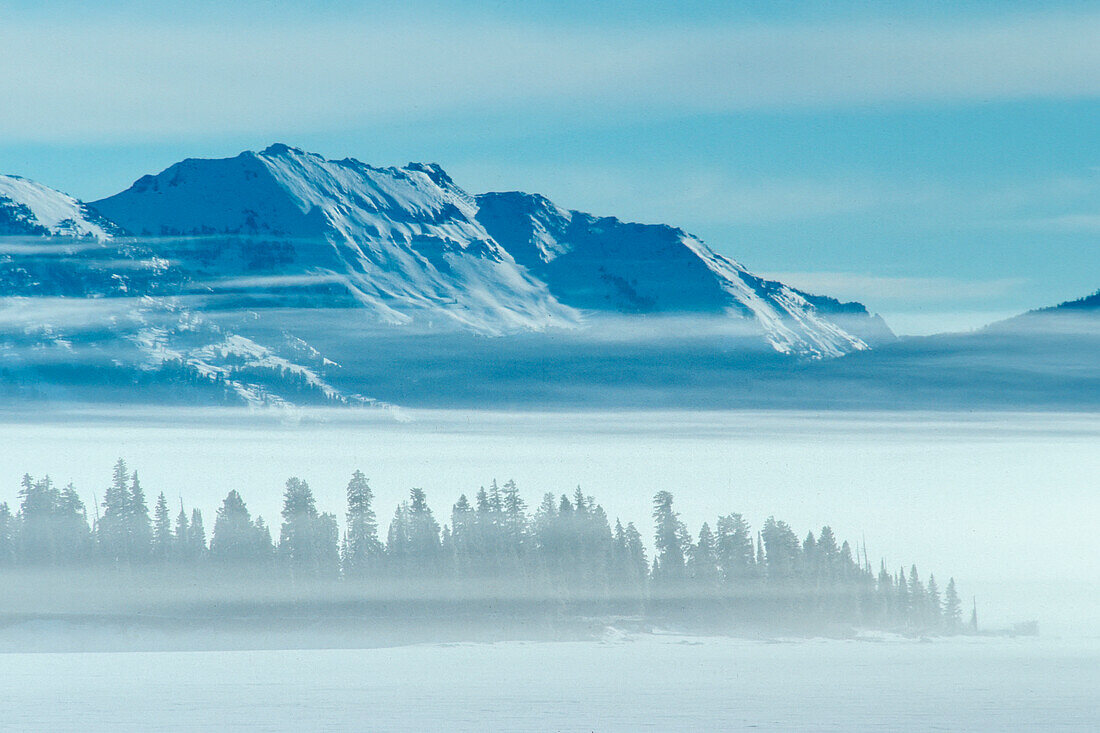 Stevenson Island, named after a member of the 1871 Hayden survey party, is the second largest of the seven islands in Yellowstone Lake. It is a low sliver of land about one mile long, seen here as a row of trees in the fog with Mount Chittenden rising ten miles beyond the island on the eastern boundary of the park in winter, Yellowstone National Park; Wyoming, United States of America