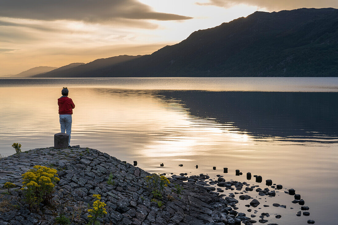 Bei Sonnenaufgang schaut ein Mann auf das ruhige Wasser des Loch Ness in Schottland; Fort Augustus, Schottland