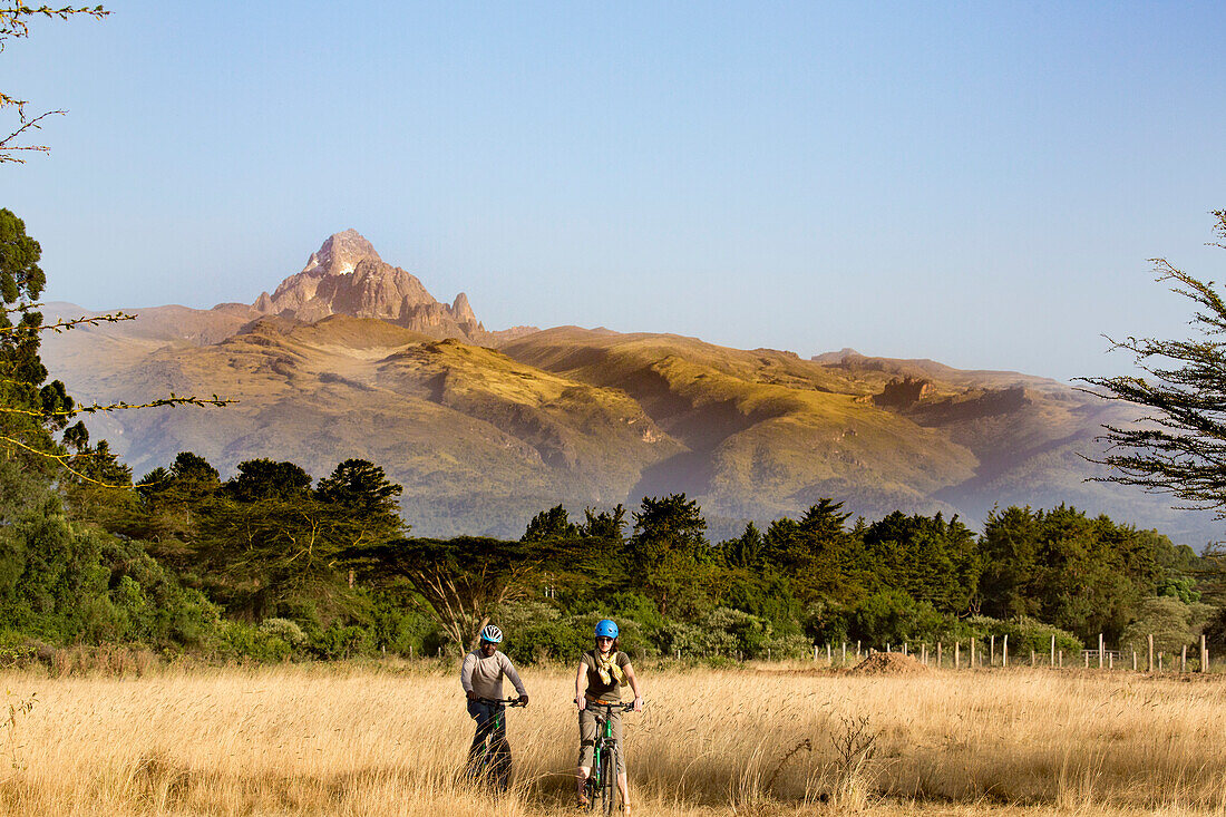 Tourist and guide on mountain bike riding safari in Mount Kenya National Park, with dramatic view of Mount Keynya with the mist rising from forest below in Nyeri County, near Nanyuki; Kenya, Africa