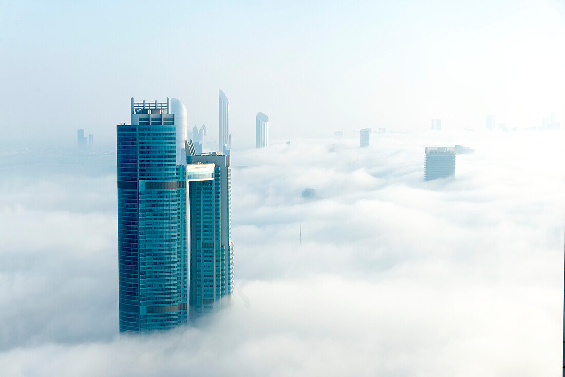 Überblick über Abu Dhabi City mit Blick auf die Nation Towers und andere Hochhäuser, darunter die beiden Türme des World Trade Center Abu Dhabi Complex in der Ferne, die durch die sonnenbeschienenen Wolken und den Nebel auftauchen; Abu Dhabi, Vereinigte Arabische Emirate