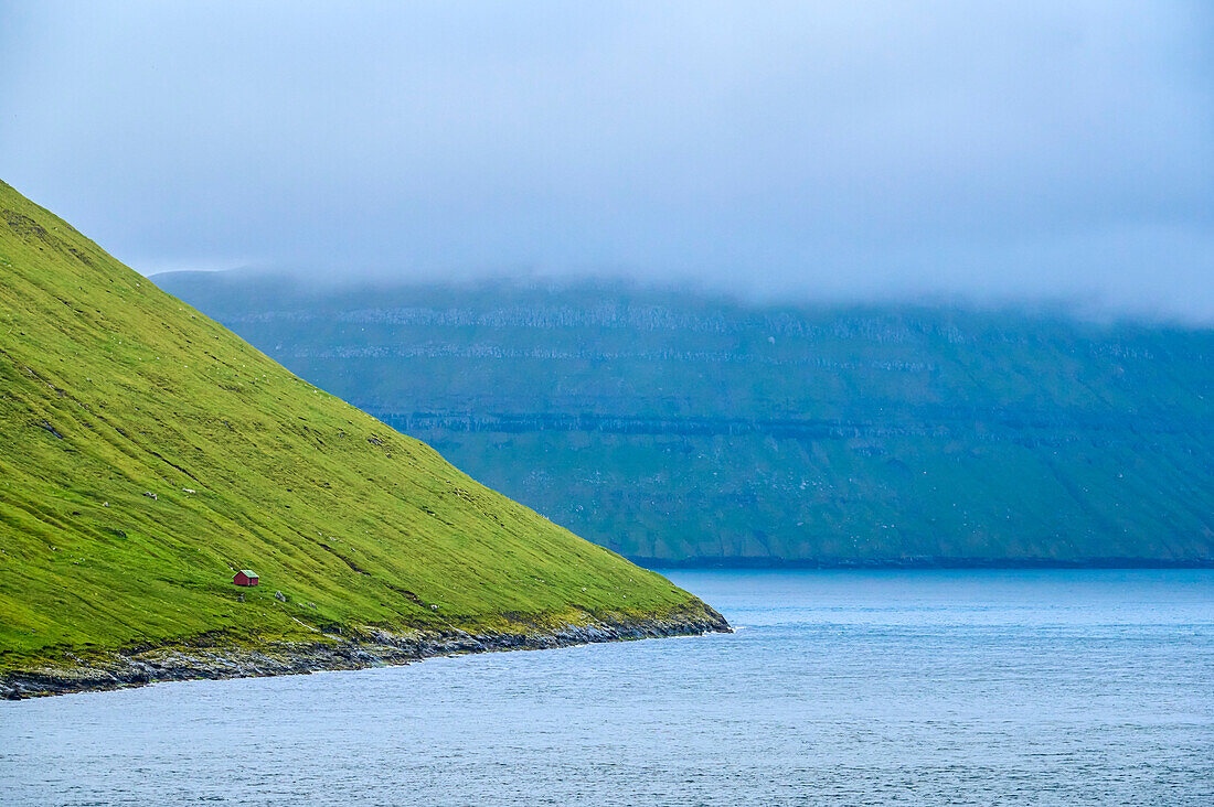 Hütte auf grasbewachsenem Berghang in der Nähe des Meeresufers, Blick von der von Island kommenden Inselfähre MS Norr?auf die Insel der Färöer, einem autonomen dänischen Territorium; Färöer Inseln
