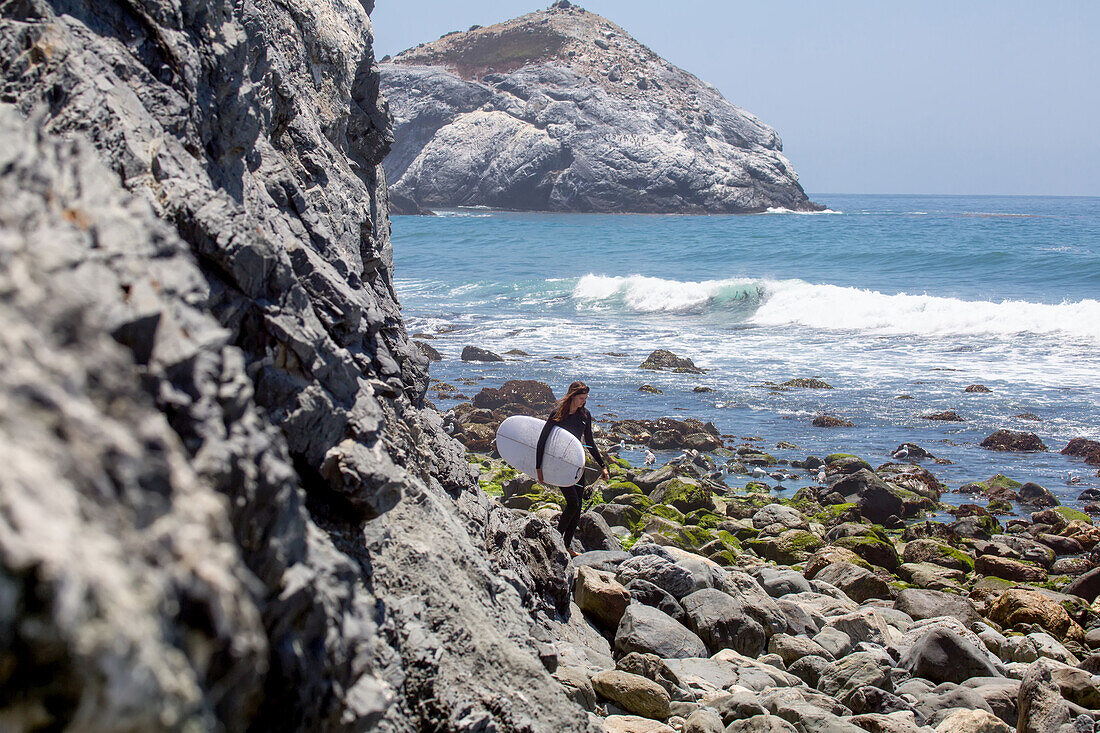 A surfer crosses a boulder strewn beach to get to the waves.