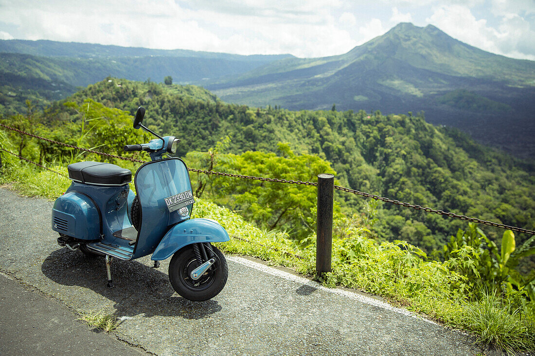 Moped parked along the side of the road at a lookout with views of Mount Batur (Kintamani Volcano) in South Batur with a cloudy sky and lush vegetation; Kintamani, Bangli Regency, Bali, Indonesia