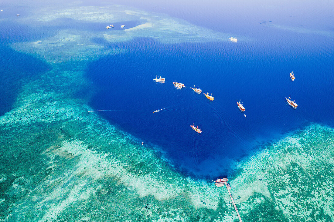 Luftaufnahme von Booten, die vor einer Insel im Komodo-Nationalpark vertäut sind, mit einem Steg, der in das umgebende türkisfarbene Wasser hineinragt; Ost Nusa Tenggara, Kleine Sunda Inseln, Indonesien