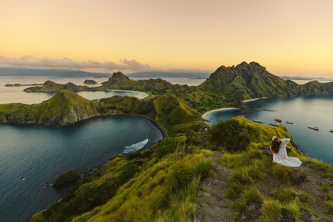 Woman sitting on the hilltop overlooking Padar Island, enjoying the sunset with  boats moored in the bay in Komodo National Park in the Komodo Archipelago; East Nusa Tenggara, Indonesia