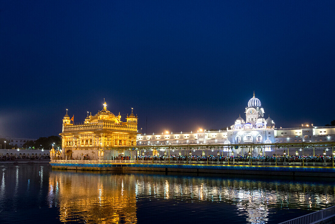 The Clock Tower and Gothic style entrance to the gilded Golden Temple (Harmandir Sahib) the most prominent holy Gurdwara Complex of the Sikh Religion with Sarovar (Sacred Pool) illuminated at night; Amritsar, Punjab, India