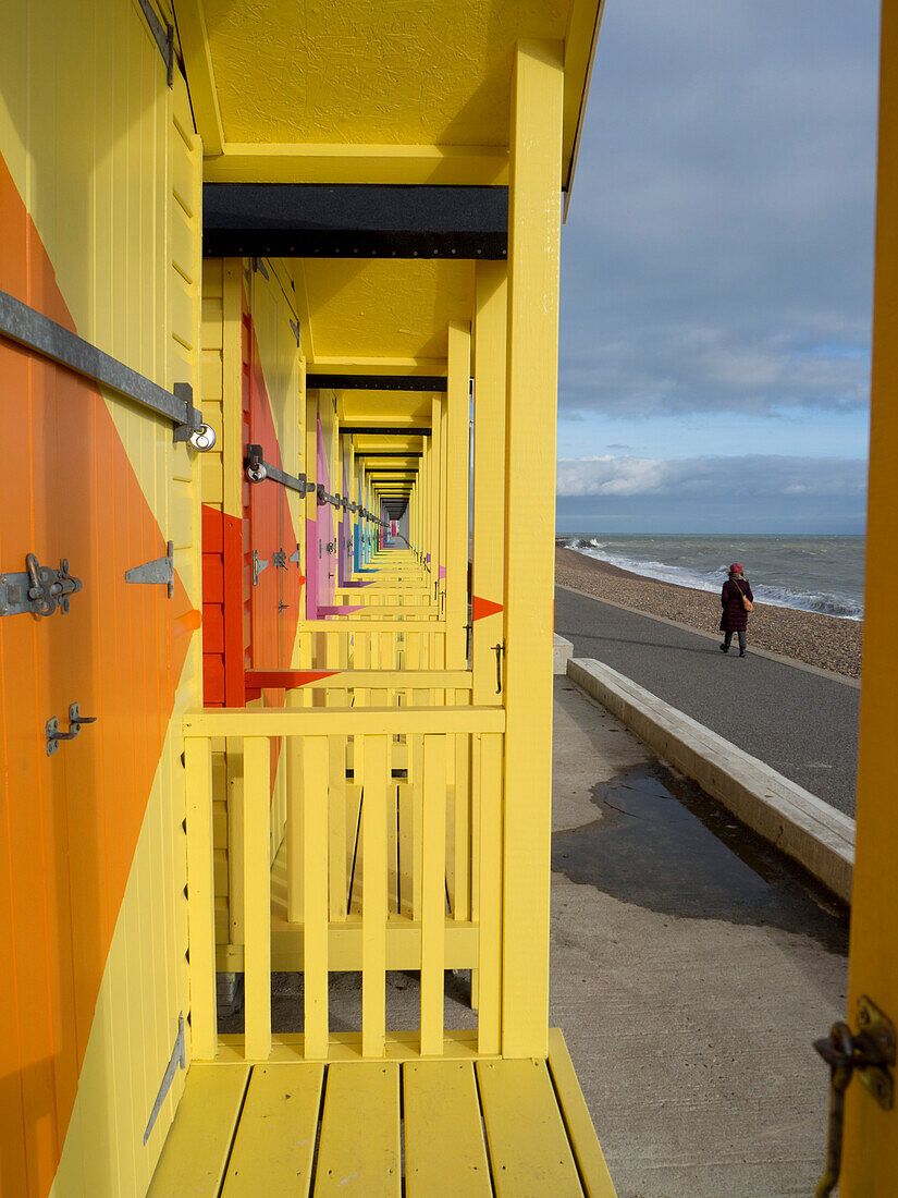 View taken from behind of a woman walking along the Atlantic shore with a one point perspective view of colorful beach huts with sea and promenade; Folkestone, Kent, England, United Kingdom