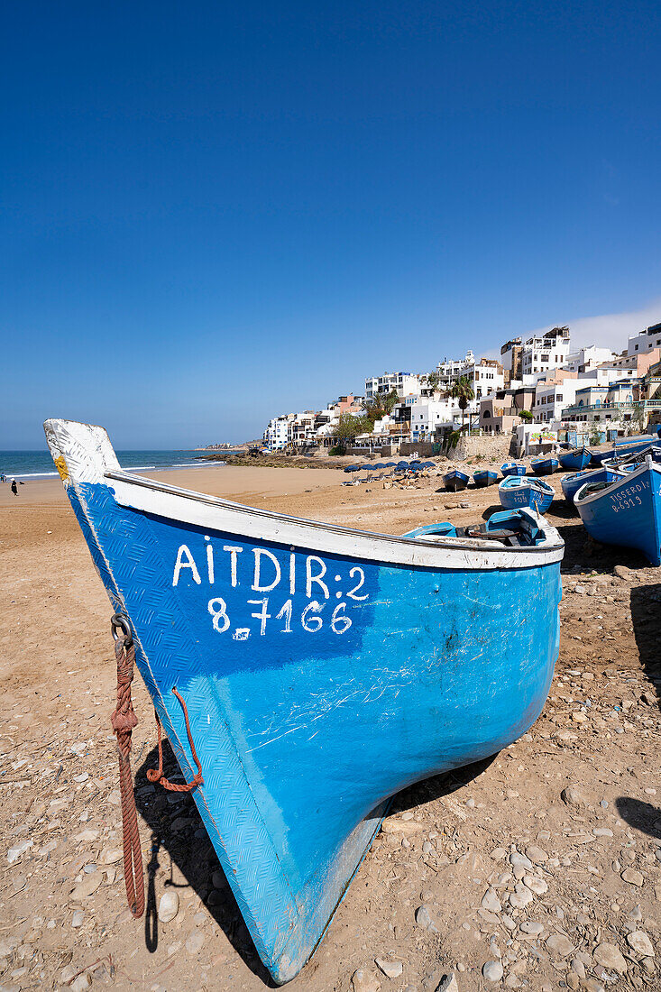 Traditionelle, leuchtend blaue Fischerboote säumen den Strand des Dorfes Taghazout mit den weißgetünchten Gebäuden am Hang im Hintergrund; Taghazout Bay, Agadir, Marokko