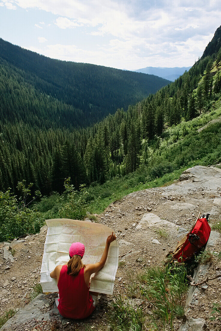 Frau prüft eine Karte am Wegesrand mit Blick auf ein baumbewachsenes Tal; Glacier National Park, Montana.
