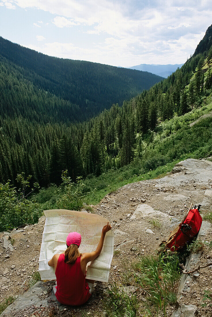 Woman examines a map trailside overlooking a tree covered valley.; Glacier National Park, Montana.