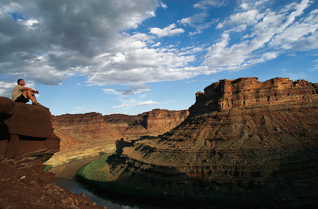 Hiker sits on cliff's edge overlooking the Colorado River.; Cataract Canyon, Utah.