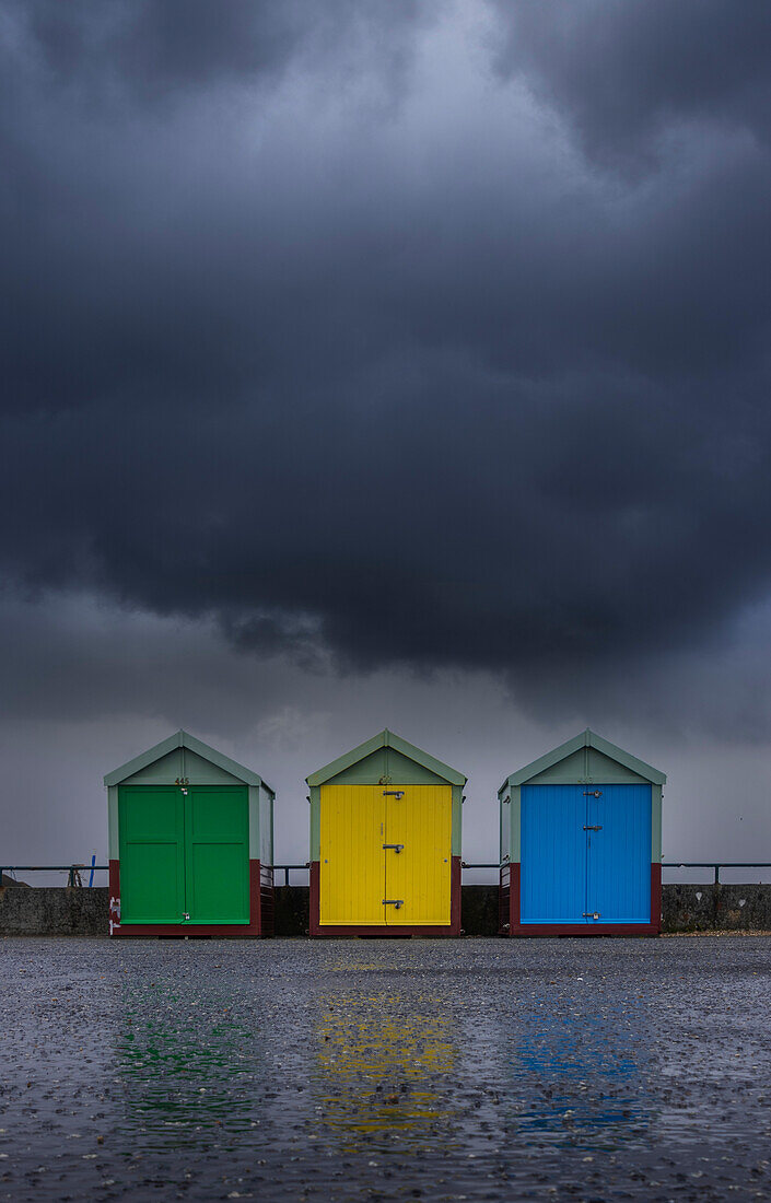 Three brightly colored beach huts sit beneath a storm cloud on a wet autumnal day; Hove, East Sussex, England, United Kingdom