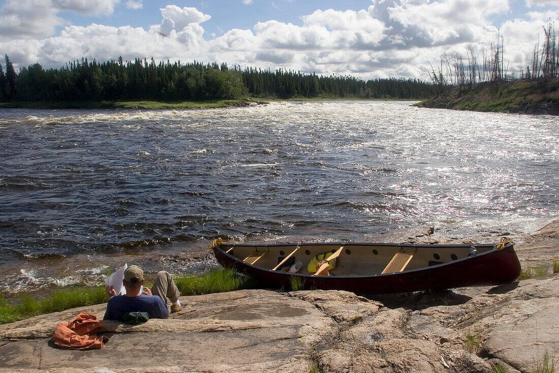 Canoeist looks at map on the shore.; Winisk River, Northern Ontario, Canada.
