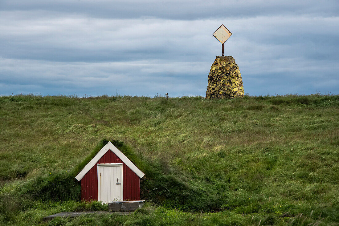 Storage structure on the side of a grassy hill and stone formation with radar reflector on the hilltop on Flatey Island, a former fishing village on the northwest side of Iceland. Part of a cluster of about forty large and small islands and islets located in Breiðafjörður. It is now a tourist town where people rent small homes in summer; Western Islands, Iceland