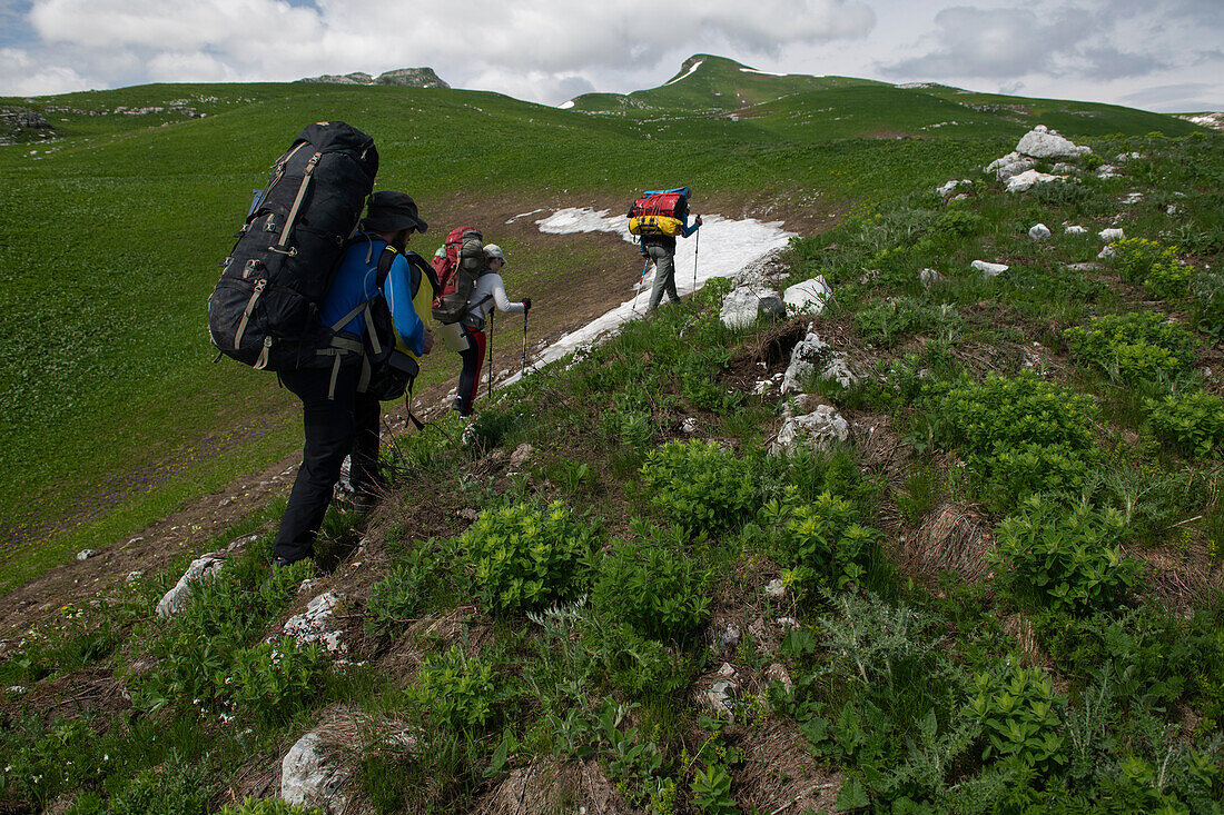 Abenteuerliche Erkundungsexpedition: Drei Forscher wandern einen grasbewachsenen Berghang hinauf und tragen ihre Ausrüstung auf dem Weg zu einem Basislager in der Nähe des Höhleneingangs von Veryovkina hoch im Kaukasusgebirge; Gagra, Kaukasusgebirge, Abchasien