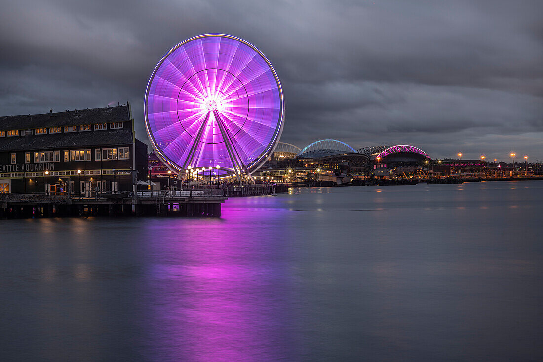 A cloudy evening on the Seattle waterfront looking at the Seattle Great Wheel reflecting off Elliott Bay with Lumen Field and T-Mobile Park lit up in the background; Seattle, Washington, United States of America