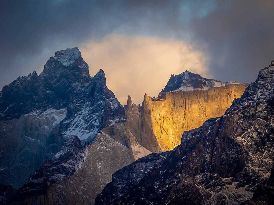 Sunlit rock faces of the jagged, mountain peaks in the Torres del Paine National Park under a stormy sky with storm light; Torres del Paine National Park, Patagonia, Chile