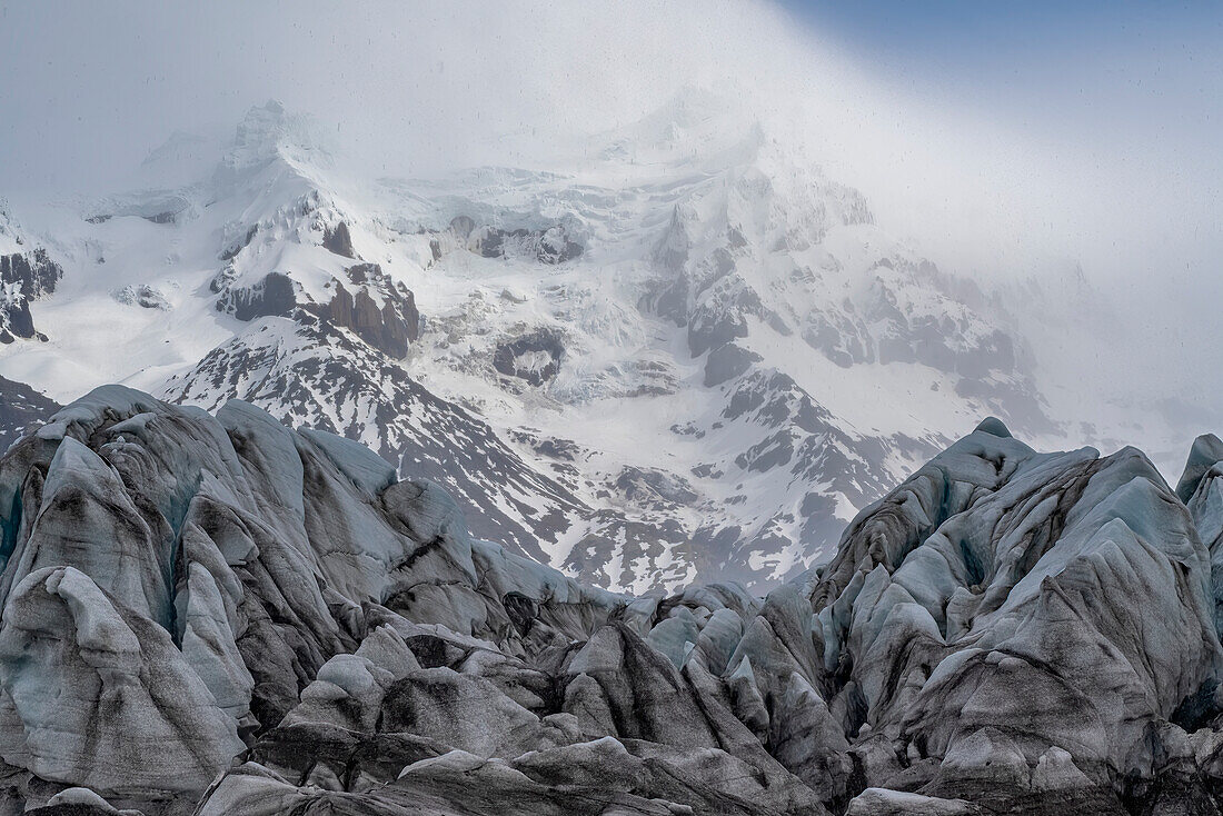 Malerischer Blick auf Gletschereisformationen und schneebedeckte Berge mit nebligen Wolken, die die Gipfel im Hintergrund einhüllen; Island