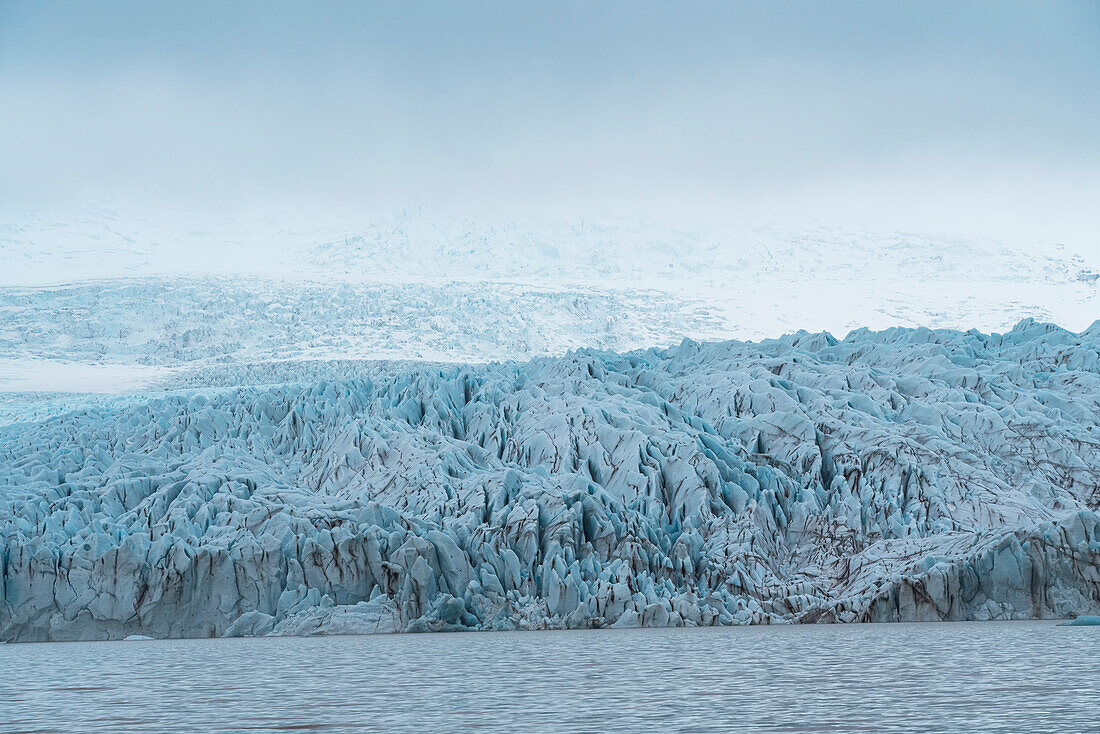 Views of the blue ice of the Fjallsjokull Glacier stands out in the misty atmosphere from the Fjallsarlon Glacier Lagoon, at the south end of the famous Icelandic glacier Vatnajökull in Southern Iceland; South Iceland, Iceland