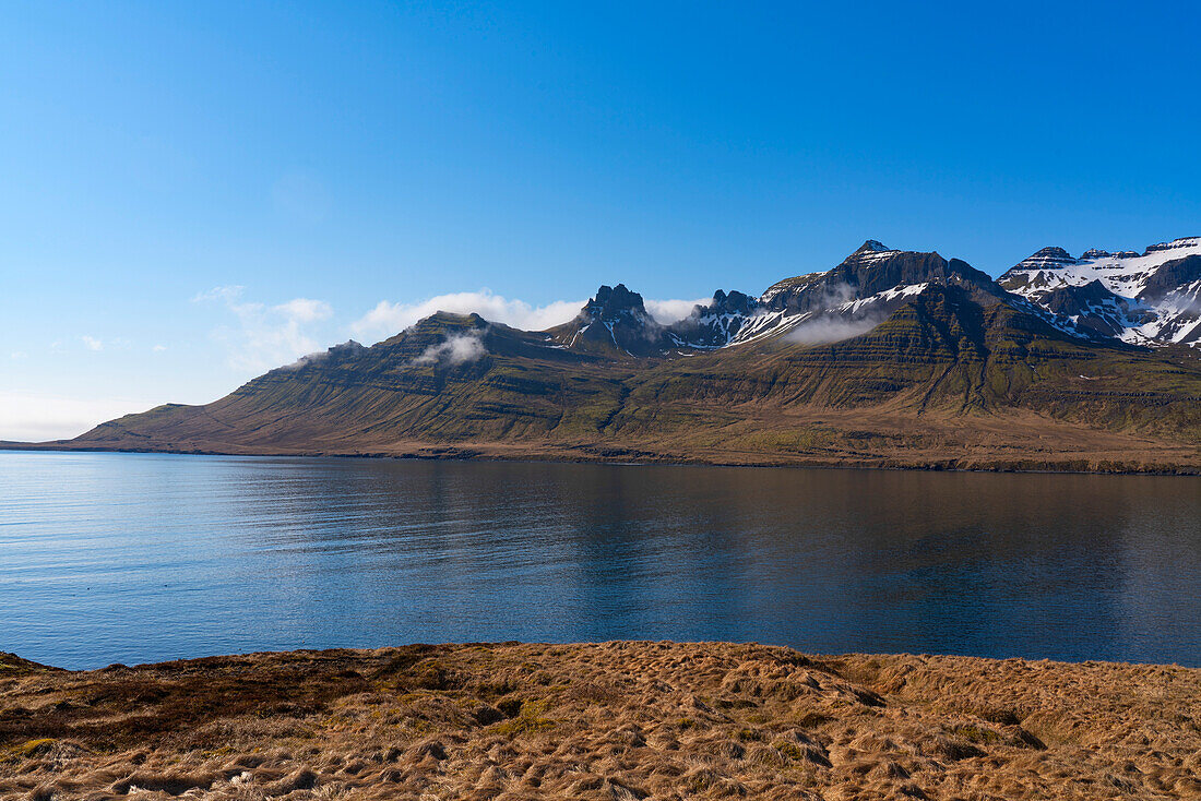 The mountainous terrain of the East Fjords with a blue sky and calm waters, creating a stunning landscape to travel through; East Iceland, Iceland