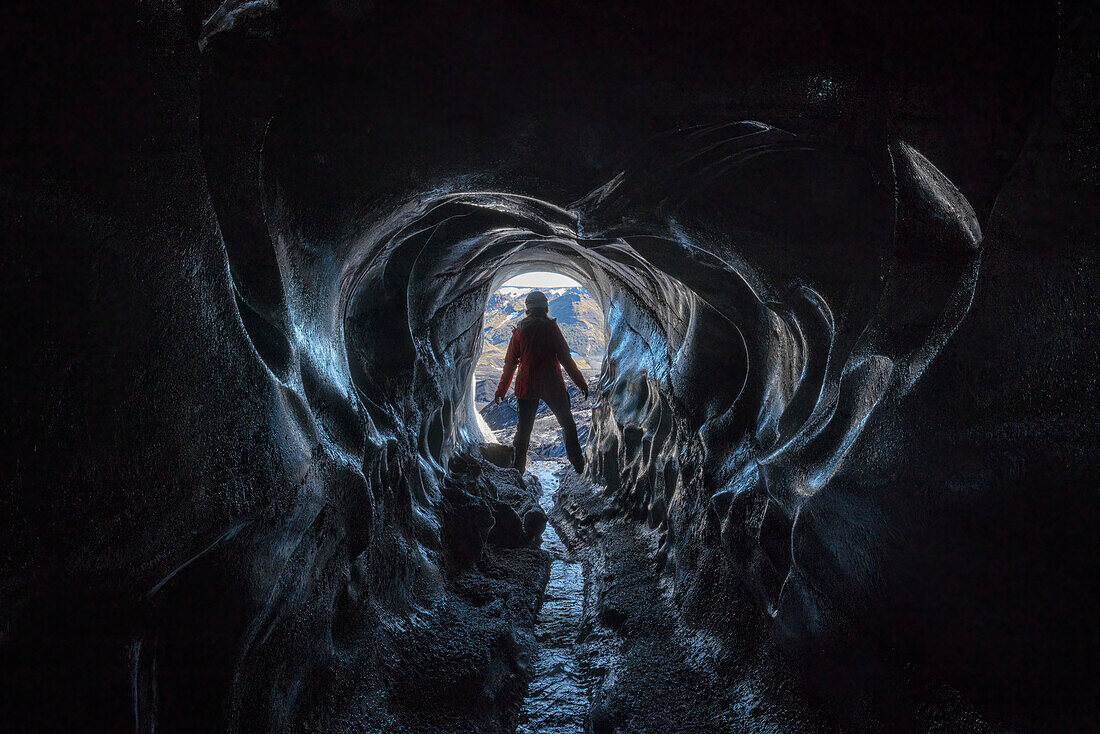 Blick durch eine Eishöhle auf eine Frau, die einen Gletscher erkundet, während sie in Island unterwegs ist; Vik, Südisland, Island