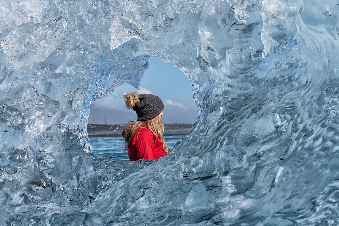 Close-up view through a round, clear ice formation of a woman wearing a woolen hat looking out and admiring the icebergs along the South Coast of Iceland; Jokulsarlon, South Iceland, Iceland
