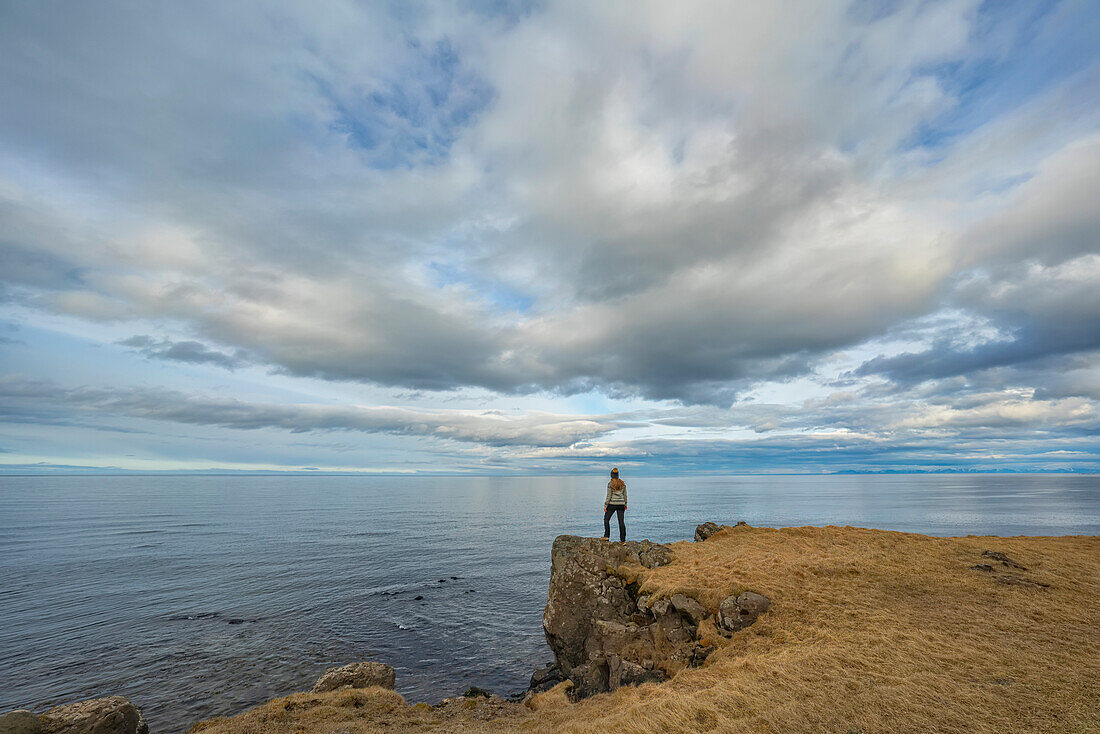 View taken from behind of a woman standing on a sea cliff overlooking the Atlantic Ocean on the North Shore of Iceland; Djupavik, Strandir Coast, Iceland