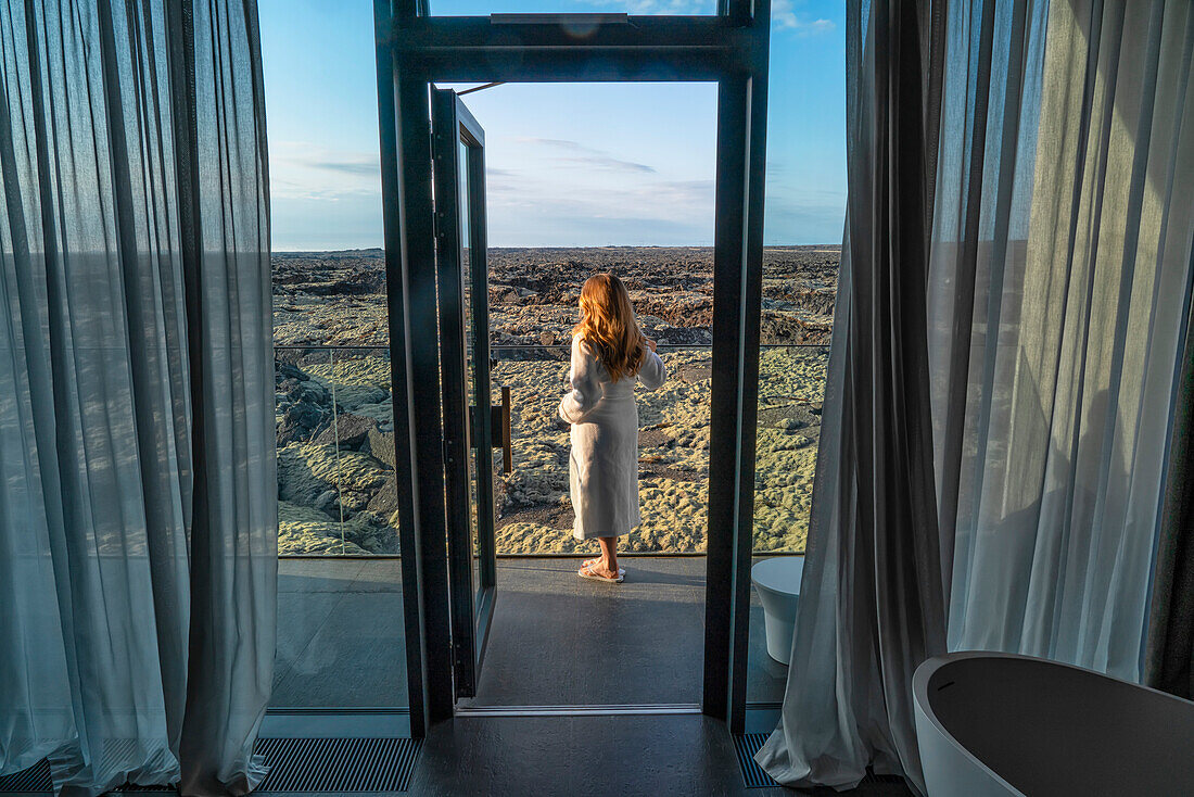 Frau steht im Bademantel auf einem Balkon mit Blick auf die Lavafelder in Südisland; Blaue Lagune, Südisland, Island
