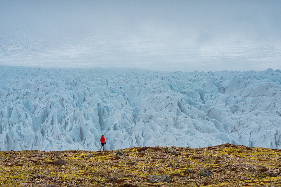 Frau geht auf der Tundra vor einem beeindruckenden Gletscher mit seinem zerklüfteten blauen Eis und der nebligen Atmosphäre entlang der Südküste Islands; Südisland, Island