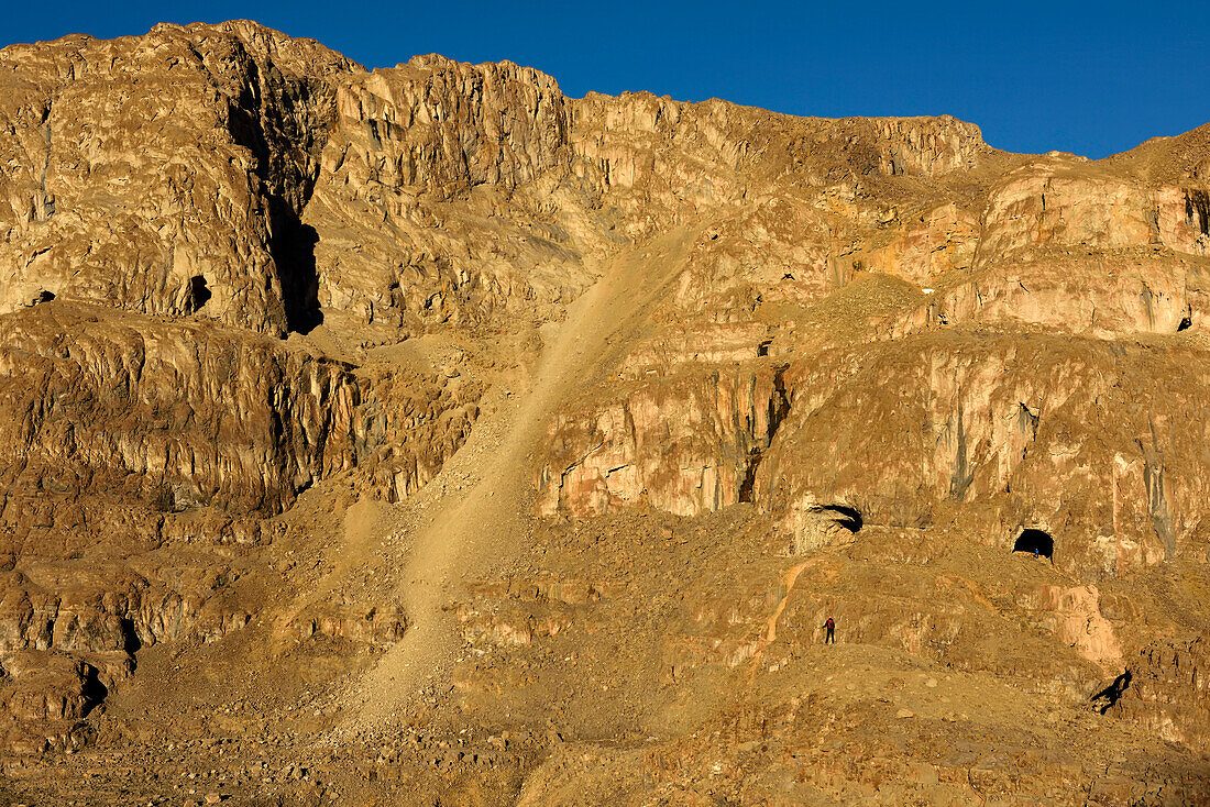 Bei Einbruch der Abendsonne steht ein Mitglied des Expeditionsteams auf einem großen Felsbrocken. In der Ferne und hinter ihm steht ein anderes Teammitglied in Blau in der Mitte des rechten Eingangs zur U-förmigen Höhle. Die riesige Geröllhalde, die größte aller Geröllhalden, die das Team auf dieser Expedition ertragen musste, fällt in der Mitte der Felswand ab und bietet den Zugang, den sie brauchten, um in den Kristallpalast zu gelangen.