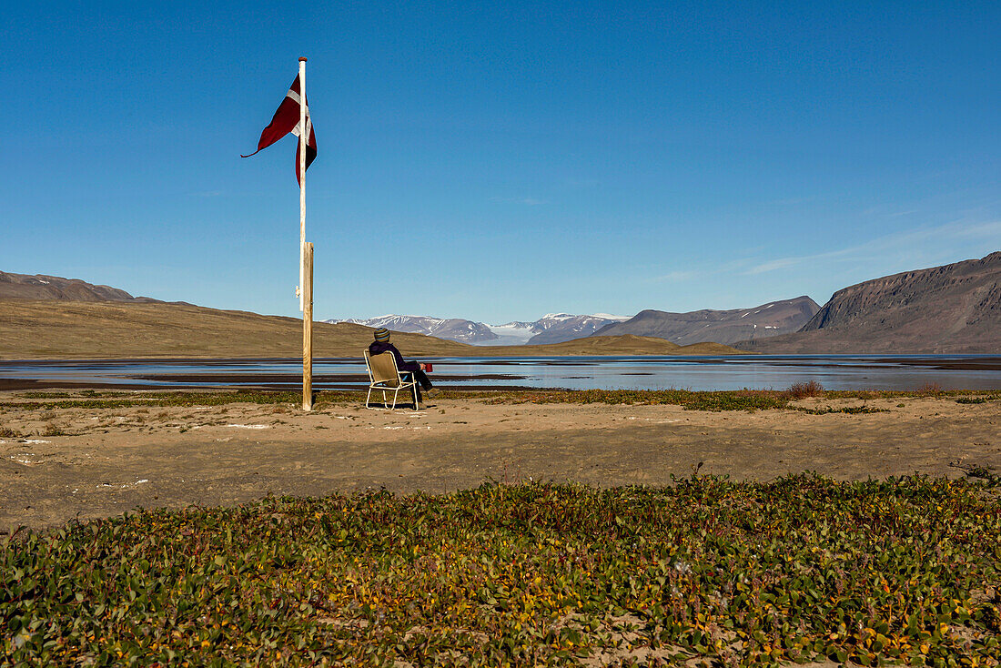 Ein Expeditionsteilnehmer sitzt auf einem alten Stuhl unter der hoch im Wind wehenden dänischen Flagge und denkt bei einer Tasse Tee über die Expedition nach. Was für ein Ort, was für ein Privileg.