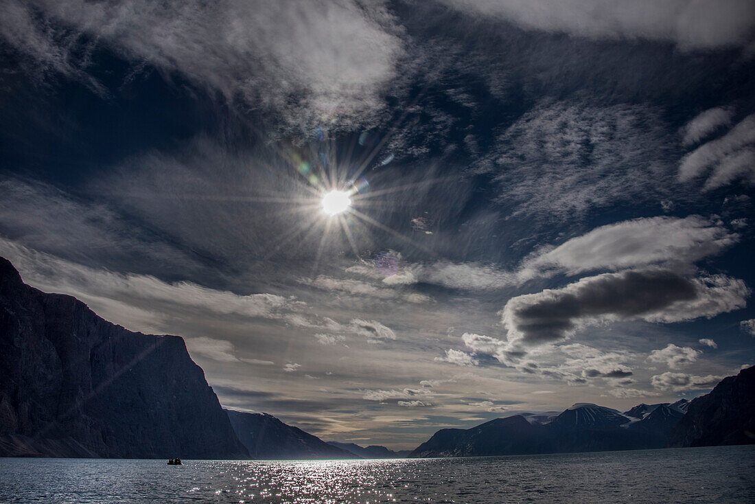 Travelers in a boat passing through Greenland's Antarctic Straits with a dramatic cloudy sky and sunburst; East Greenland, Greenland