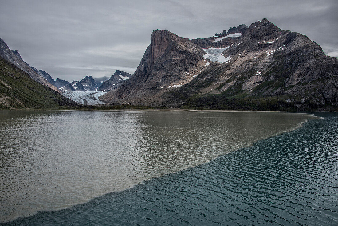 Blick auf einen Gletscher, der durch die Berggipfel an der Südspitze Grönlands fließt, mit zwei Wassertypen, die sich am Gletschersockel in Prins Christian Sund unter einem grauen Himmel vermischen; Südgrönland, Grönland